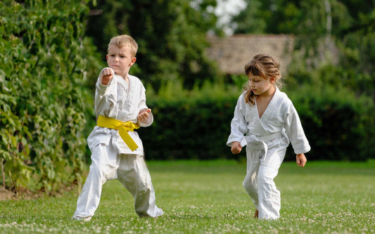 Two children in white martial arts uniforms practicing outdoors on grass, with one performing a punch and the other observing, surrounded by greenery.