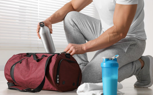 A person in athletic wear is packing a maroon gym bag with a silver water bottle. The person is kneeling on the floor, and there is a blue shaker bottle and white towel next to the gym bag