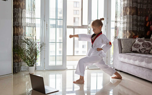 Child in a white martial arts uniform practicing punches while following an online class on a laptop in a bright living room.