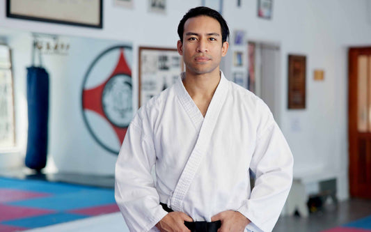 Person in a white martial arts uniform standing in a dojo, hands resting on their belt, with mats, a punching bag, framed pictures, and a circular red-and-white emblem in the background