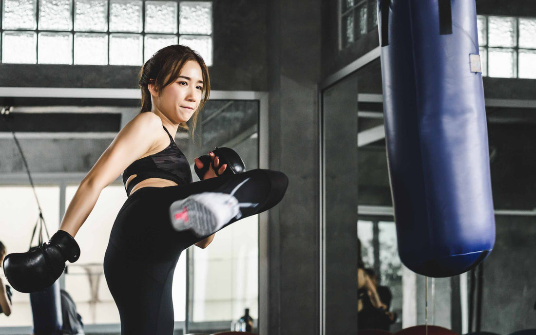 Person in athletic gear delivering a high kick to a blue punching bag in a well-lit gym with glass block windows and mirrors