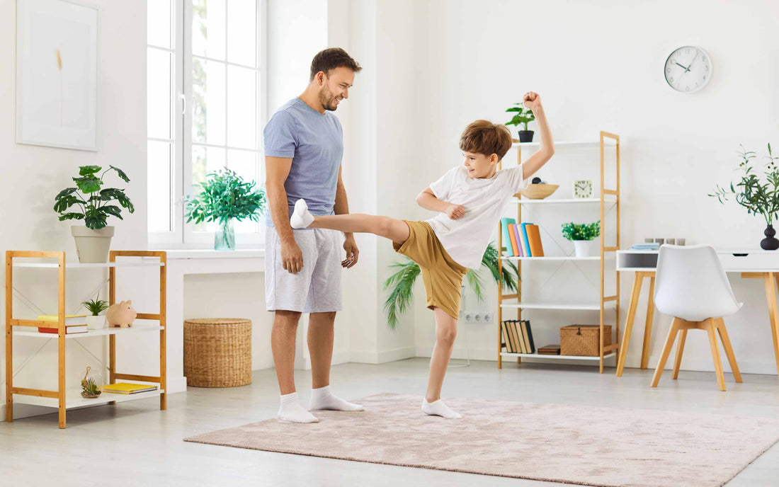 A smiling father watching his young son practice a martial arts high kick in their living room, demonstrating an at-home training routine.