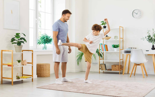 A smiling father watching his young son practice a martial arts high kick in their living room, demonstrating an at-home training routine.